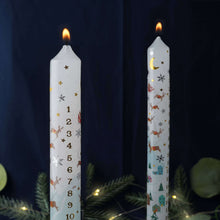 Load image into Gallery viewer, White Christmas advent candle decorated with gold stars and snowflakes, lit wick glowing, set against dark background