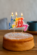 Load image into Gallery viewer, Lit rainbow 'Happy Birthday' candle on a sponge cake dusted with icing sugar and colourful sprinkles, displayed on a wooden table with a blurred mug and cushion in the background