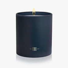 Load image into Gallery viewer, A dark blue candle in a matching cylindrical container, lit with a visible flame and featuring a gold Roman numeral III logo on the front, set against a clean white background.
