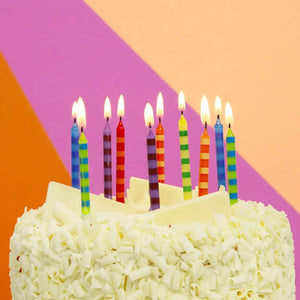 Lit striped rainbow birthday candles on a white chocolate cake against a vibrant orange, pink, and purple background – fun and festive cake decoration for parties